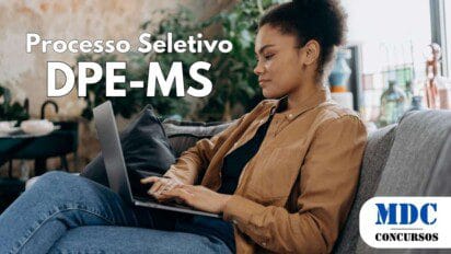 Mulher jovem de cabelos cacheados presos em um coque volumoso, vestindo camisa marrom e calça jeans, sentada confortavelmente em um sofá cinza com almofadas, usando um laptop. O ambiente tem plantas e decoração moderna, com luz natural entrando pela janela - Processo Seletivo DPE-MS em Salário de R$ 5.325,59 em Cadastro de Reserva