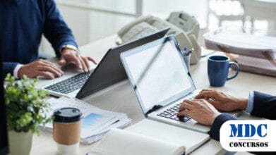 Dois profissionais em trajes formais trabalham em laptops sobre uma mesa de escritório, cercados por papéis, cadernos, café e itens de escritório. A tela de um dos laptops exibe gráficos. O ambiente é moderno e bem iluminado, com telefone e xícara ao fundo, sugerindo um ambiente corporativo colaborativo / Processo Seletivo Piauí 2025