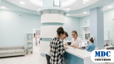 Recepção de uma clínica com ambiente moderno em tons de azul e branco. Uma funcionária atende duas pessoas no balcão, enquanto outra trabalha no computador. Ao fundo, duas mulheres conversam perto de uma estante com produtos. O local tem sofás, iluminação suave e um design limpo e organizado - Processo Seletivo SES SC 2025 tem 23 Vagas e Remuneração de até R$ 10,2 mil