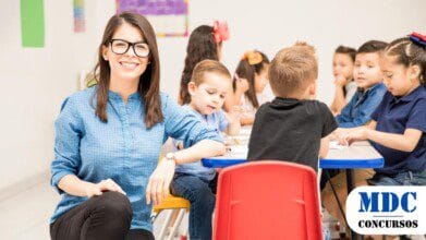 Professora sorridente com óculos e blusa azul em sala de aula com crianças sentadas em mesas coloridas, envolvidas em atividades. O ambiente é iluminado e decorado com materiais educativos. - Processo Seletivo Semed Manaus 2025