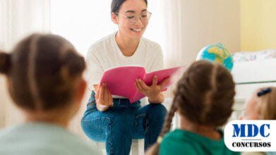 Processos Seletivos 6 Mulher jovem de óculos e camiseta branca lê um livro rosa para três crianças sentadas à sua frente. As crianças, de cabelos trançados e amarrados, ouvem atentamente. O ambiente é iluminado, com um globo terrestre e móveis brancos ao fundo, sugerindo um espaço educativo ou infantil - Processo Seletivo em Anápolis - GO oferece 350 vagas com remuneração de até R$ 2 mil
