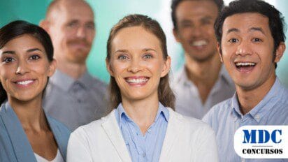 Grupo de cinco pessoas sorrindo em um ambiente corporativo. A mulher ao centro, de blazer branco, se destaca na imagem, cercada por colegas com expressões alegres. O fundo é esverdeado e desfocado, enfatizando os rostos iluminados. Todos vestem roupas formais ou semiformais, transmitindo um clima profissional e positivo / Processo Seletivo em Assis Chateaubriand oferece salários de até R$16,6 mil