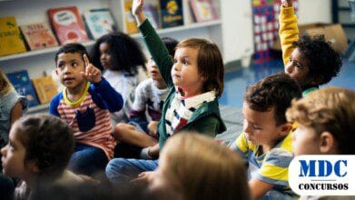 Processos Seletivos 7 Crianças sentadas no chão de uma sala de aula, participando ativamente da atividade. Algumas levantam as mãos, demonstrando interesse. Ao fundo, há prateleiras com livros coloridos / Processo Seletivo na Educação de Bujari – AC Abre 68 Vagas com Remuneração de R$ 2,9 mil