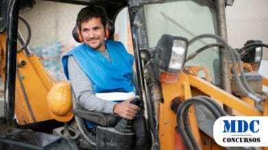 Operador de retroescavadeira usando colete azul, protetores auriculares e roupa cinza, sentado na cabine da máquina segurando o controle. Um capacete amarelo está apoiado ao lado. O cenário mostra um ambiente de construção com materiais ao fundo/Realeza (PR) Oferece Salários de Até R$ 3 mil em Processo Seletivo