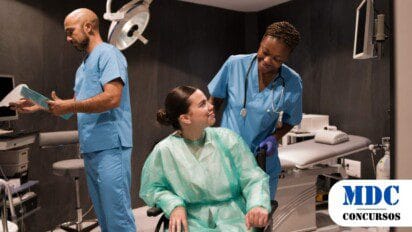 Equipe médica em ambiente hospitalar. Enfermeira sorri para paciente em cadeira de rodas, que veste avental descartável. Médico de avental azul segura prontuário e analisa documentos. Sala com equipamentos médicos, maca, monitor e cilindro de oxigênio. Iluminação clínica e atmosfera profissional - Resultado Final do Processo Seletivo 2025 na Saúde de Cruzeiro do Sul é Publicado