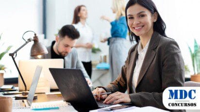 Mulher sorridente de terno marrom trabalha em um laptop em um escritório moderno. Ao fundo, um homem trabalha concentrado e duas mulheres conversam. A mesa contém cadernos, caneta, celular e luminária. O ambiente é bem iluminado e profissional / SAAE Passos MG Abre Processo Seletivo com 5 Vagas + Cadastro de Reserva