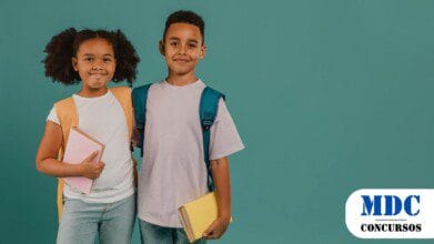 Duas crianças sorridentes vestindo mochilas e segurando livros, posam juntas contra um fundo verde. A menina usa uma mochila laranja e segura um livro rosa, enquanto o menino tem uma mochila azul e segura um livro amarelo. Eles vestem roupas casuais e parecem prontos para a escola - SEDUC Juazeiro (BA) lança Processo Seletivo para CR com Salários de Até R$ 2,5 mil