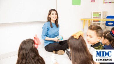 Processos Seletivos 2 Professora sentada no chão de uma sala de aula, sorrindo e segurando uma bola azul, enquanto interage com crianças ao seu redor. As crianças, com laços coloridos no cabelo, estão sentadas em círculo e parecem atentas. O ambiente é claro, com cartazes educativos na parede, uma lousa branca e móveis escolares ao fundo / SEDUC convoca mais de 800 profissionais para suprir demandas da Rede Estadual de Ensino do Amazonas