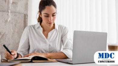 Processos Seletivos 9 Mulher de camisa branca trabalhando em um laptop, escrevendo em um caderno com uma caneta. Ela está concentrada, sentada em uma mesa com um café ao lado. O ambiente é moderno, com luz natural entrando por uma janela com persianas brancas - SEJURI SC Abre novo Processo Seletivo