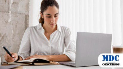 Mulher de camisa branca trabalhando em um laptop, escrevendo em um caderno com uma caneta. Ela está concentrada, sentada em uma mesa com um café ao lado. O ambiente é moderno, com luz natural entrando por uma janela com persianas brancas - SEJURI SC Abre novo Processo Seletivo
