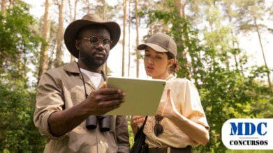 Processos Seletivos 6 Dois pesquisadores ao ar livre, em uma floresta, analisam um tablet. O homem usa chapéu, óculos e binóculos no pescoço, enquanto a mulher veste boné e uniforme bege. Ambos parecem discutir informações na tela. O ambiente é verde e iluminado pelo sol, com árvores e vegetação ao fundo / SEMA-MT Abre Processo Seletivo