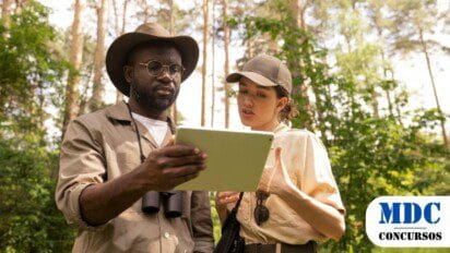 Dois pesquisadores ao ar livre, em uma floresta, analisam um tablet. O homem usa chapéu, óculos e binóculos no pescoço, enquanto a mulher veste boné e uniforme bege. Ambos parecem discutir informações na tela. O ambiente é verde e iluminado pelo sol, com árvores e vegetação ao fundo / SEMA-MT Abre Processo Seletivo
