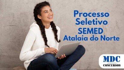 Jovem sorridente com cabelos cacheados em tranças, vestindo moletom branco, calça jeans e meias listradas, sentada no chão com as pernas cruzadas sobre uma almofada, segurando um laptop prata, olhando para o lado contra um fundo de parede cinza - SEMED Atalaia do Norte (AM) Lança Processo Seletivo 2025