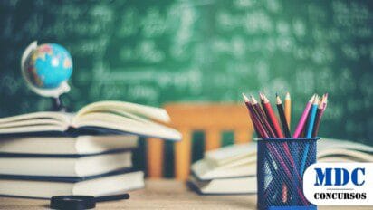 Mesa de madeira com lápis de cor em um porta-lápis azul, livros abertos e uma lupa. Ao fundo, um quadro-negro com fórmulas matemáticas escritas e um globo terrestre. Cena representa um ambiente escolar e de estudo / SEMED Maceió
