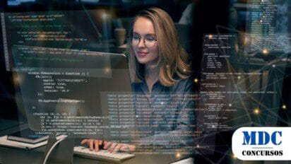 Mulher com óculos e cabelo loiro trabalha em um computador em um ambiente moderno. Códigos e elementos gráficos futuristas aparecem sobrepostos, representando tecnologia e programação - SESI/SENAI Amapá Lançam Processo Seletivo para Analista de TI