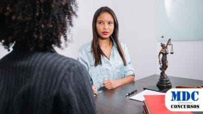Duas mulheres conversam em um escritório jurídico. A mulher de frente usa uma blusa azul e parece ouvir atentamente. Na mesa, há livros, documentos, um martelo de juiz e uma estátua da Justiça. O ambiente é profissional, com um quadro de vidro ao fundo / STM lança Concurso Público com 80 vagas e remuneração de até R$ 14,8 mil