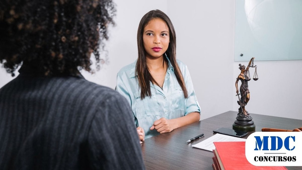 STM lança Concurso Público com 80 vagas e remuneração de até R$ 14,8 mil 1 Duas mulheres conversam em um escritório jurídico. A mulher de frente usa uma blusa azul e parece ouvir atentamente. Na mesa, há livros, documentos, um martelo de juiz e uma estátua da Justiça. O ambiente é profissional, com um quadro de vidro ao fundo / STM lança Concurso Público com 80 vagas e remuneração de até R$ 14,8 mil