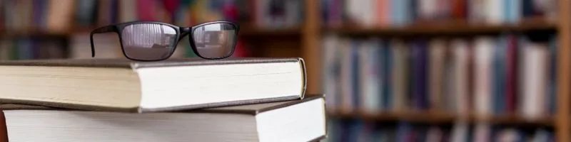 São Luís Abre 146 Vagas em Processo Seletivo no Combate às Endemias 2 Pilha de livros com capa dura em uma biblioteca, com um par de óculos de grau apoiado no topo. Ao fundo, estantes repletas de livros estão levemente desfocadas, criando um ambiente de estudo e leitura - São Luís Abre 146 Vagas em Processo Seletivo no Combate às Endemias (foto: Freepik)