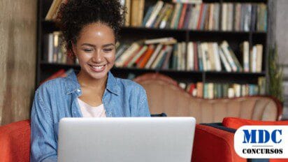 Mulher jovem de cabelo cacheado e pele morena sorri enquanto usa um laptop. Ela está sentada em um sofá vermelho, vestindo uma camisa jeans. Ao fundo, há uma estante cheia de livros. O ambiente tem um clima aconchegante e parece ser uma biblioteca ou café - São Luís Abre 146 Vagas em Processo Seletivo no Combate às Endemias