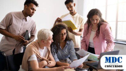 Grupo diverso de cinco pessoas em reunião, analisando documentos e usando um laptop. Duas mulheres sentadas examinam papéis, enquanto três colegas em pé observam e seguram pastas. A equipe parece concentrada e engajada, trabalhando em um ambiente iluminado por luz natural, com materiais de escritório espalhados sobre a mesa - São Miguel do Guaporé (RO) convoca 74 Aprovados em Processo Seletivo