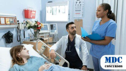 Médico e enfermeira conversam ao lado de uma paciente deitada em um leito hospitalar. O médico veste jaleco branco e estetoscópio, enquanto a enfermeira segura uma pasta azul. Equipamentos médicos, um buquê de flores e uma tabela de visão estão ao fundo, em um ambiente hospitalar iluminado - SES PE Oferece 259 Vagas com Salários de até R$ 11,5 mil em Processo Seletivo