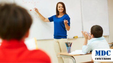 Processos Seletivos 3 Professora sorridente de blusa azul escreve equações no quadro branco enquanto alunos, sentados em carteiras escolares, participam da aula. Um aluno de camisa branca levanta a mão, enquanto outro, de vermelho, observa. O ambiente é iluminado e organizado, com materiais escolares sobre as mesas / Seletivo da Secretaria de Educação de Pernambuco Oferece 338 Vagas