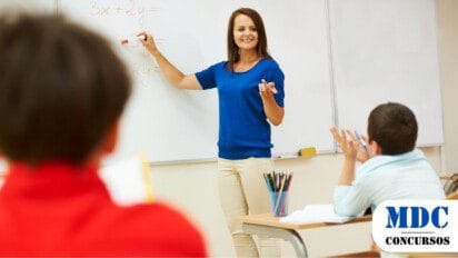 Professora sorridente de blusa azul escreve equações no quadro branco enquanto alunos, sentados em carteiras escolares, participam da aula. Um aluno de camisa branca levanta a mão, enquanto outro, de vermelho, observa. O ambiente é iluminado e organizado, com materiais escolares sobre as mesas / Seletivo da Secretaria de Educação de Pernambuco Oferece 338 Vagas