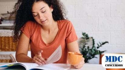 Mulher de cabelos cacheados e pele clara veste camiseta laranja e está sentada à mesa, segurando uma xícara enquanto lê um livro ou revista. O ambiente tem decoração aconchegante, com plantas e cestos organizadores ao fundo. Ela parece concentrada na leitura, em um espaço bem iluminado e tranquilo / Seletivo em Cotriguaçu (MT) Oferece Salários de Até R$ 5 Mil