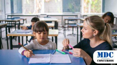 Professora auxiliando uma aluna em atividade com lápis e canetas coloridas em sala de aula, enquanto outras crianças estudam ao fundo / Semed Altos (PI) Convoca Classificados na Primeira Chamada do Edital 001/2025