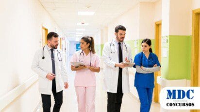 Profissionais da saúde caminhando em um corredor de hospital, discutindo informações em tablets. Dois médicos usam jaleco branco e estetoscópio, enquanto duas enfermeiras vestem uniformes rosa e azul. Eles parecem concentrados e engajados na conversa, demonstrando trabalho em equipe e colaboração - Sesap RN Convoca 78 Aprovados em Processo Seletivo