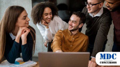 Grupo de cinco jovens profissionais reunidos ao redor de um laptop, sorrindo e interagindo de forma animada em um ambiente de escritório moderno - TCE SP Abre Concurso com 63 Vagas e Salários de até R$ 17,7 mil
