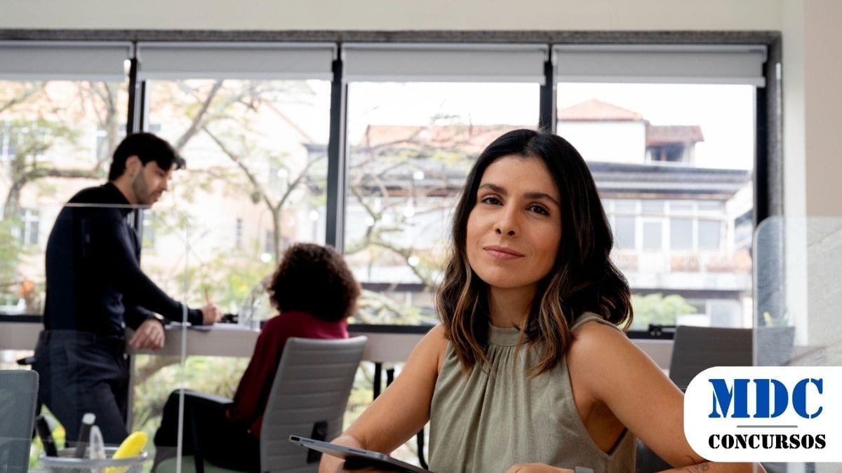 TJRR Concurso 2025 para Serventias Extrajudiciais Abre Vagas para Notários 1 Mulher com cabelo castanho ondulado, vestindo blusa verde e smartwatch, segura um tablet e sorri levemente para a câmera em um escritório moderno. Ao fundo, duas pessoas conversam próximas a uma janela grande com vista para árvores e prédios. O ambiente é iluminado e profissional / TJRR Concurso para Serventias Extrajudiciais Abre Vagas para Notários e Registradores