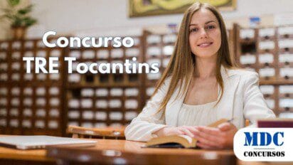 Mulher sorridente de cabelos longos e loiros, vestindo blazer branco, sentada em uma biblioteca clássica com fichários de madeira ao fundo, segurando um livro aberto sobre a mesa - TRE TO Lança Concurso com Salários de Até R$ 14,9 Mil