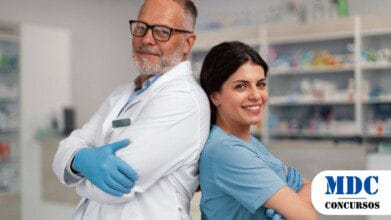 Dois profissionais de saúde, um homem mais velho de jaleco branco e uma mulher mais jovem de uniforme azul, posam sorrindo de costas um para o outro em uma farmácia. Ambos usam luvas azuis e transmitem confiança e profissionalismo. Prateleiras com produtos farmacêuticos estão ao fundo / Theobroma - RO lança Processo Seletivo 2025