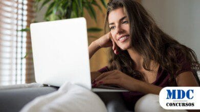 Mulher jovem de cabelos longos e castanhos, sorrindo enquanto usa um laptop. Ela está relaxada, apoiando a cabeça em uma das mãos, com unhas pintadas de vermelho. Usa uma blusa roxa e calça branca, sentada em um sofá com almofadas, com uma planta verde ao fundo - UEPB Lança 2 Processos Seletivos com Salários de até R$ 3.566,25