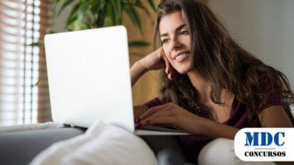 Mulher jovem de cabelos longos e castanhos, sorrindo enquanto usa um laptop. Ela está relaxada, apoiando a cabeça em uma das mãos, com unhas pintadas de vermelho. Usa uma blusa roxa e calça branca, sentada em um sofá com almofadas, com uma planta verde ao fundo - UEPB Lança 2 Processos Seletivos com Salários de até R$ 3.566,25