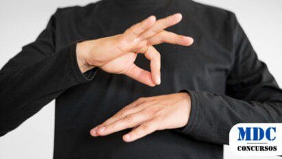 Pessoa vestindo roupa preta fazendo um sinal em Língua de Sinais com as mãos. A mão dominante forma um círculo com o polegar e o indicador, enquanto os outros dedos estão estendidos. A outra mão está aberta e posicionada horizontalmente abaixo. Fundo branco neutro - UFAL Abre Processo Seletivo