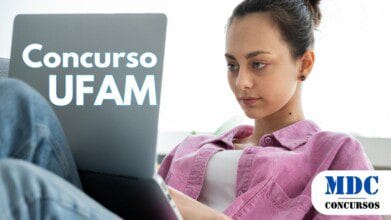 Jovem de cabelos presos usa uma camisa rosa e jeans enquanto mexe em um notebook, sentada confortavelmente em um ambiente iluminado com luz natural - UFAM Abre 74 Vagas com Salários de Até R$ 10.481,64 em Concurso Público