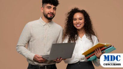 Dois jovens sorridentes, um homem com laptop e uma mulher com livros e pastas, posam contra um fundo bege. Ambos vestem roupas sociais elegantes, transmitindo um ar profissional e acadêmico / UFRGS Abre Processo Seletivo para Projeto Mais Saúde com Agente
