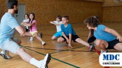 Grupo de crianças alongando-se com um professor de educação física em uma quadra coberta. Elas sorriem enquanto realizam um exercício de flexibilidade, vestindo roupas esportivas coloridas. O professor, de camiseta azul e bermuda cinza, demonstra o movimento. O ambiente tem piso de madeira e paredes revestidas de madeira compensada, com uma cesta de basquete ao fundo - Urucânia (MG) Lança Processo Seletivo para Professor de Educação Física com Salário de R$ 3 mil