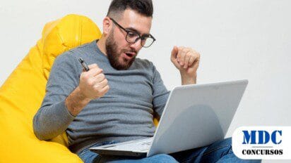 Homem de óculos e barba, sentado em uma poltrona amarela, usando um notebook. Ele segura uma caneta e demonstra expressão de alegria e comemoração. No canto inferior direito, há um logotipo com o texto "MDC Concursos" em azul e preto - Videira (SC) Lança Processos Seletivos com Remuneração de até R$ 25,1 mil