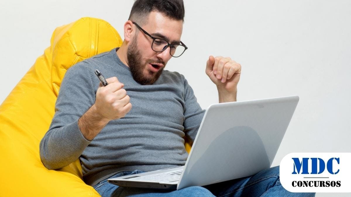 Homem de óculos e barba, sentado em uma poltrona amarela, usando um notebook. Ele segura uma caneta e demonstra expressão de alegria e comemoração. No canto inferior direito, há um logotipo com o texto "MDC Concursos" em azul e preto - Videira (SC) Lança Processos Seletivos com Remuneração de até R$ 25,1 mil