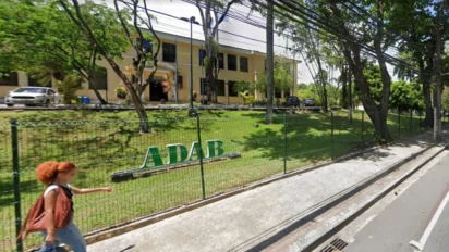 Fachada da sede da ADAB com prédio amarelo ao fundo, área arborizada, grade com logotipo ADAB e calçada com pedestre passando em frente ao local. Foto: Divulgação/Google Maps