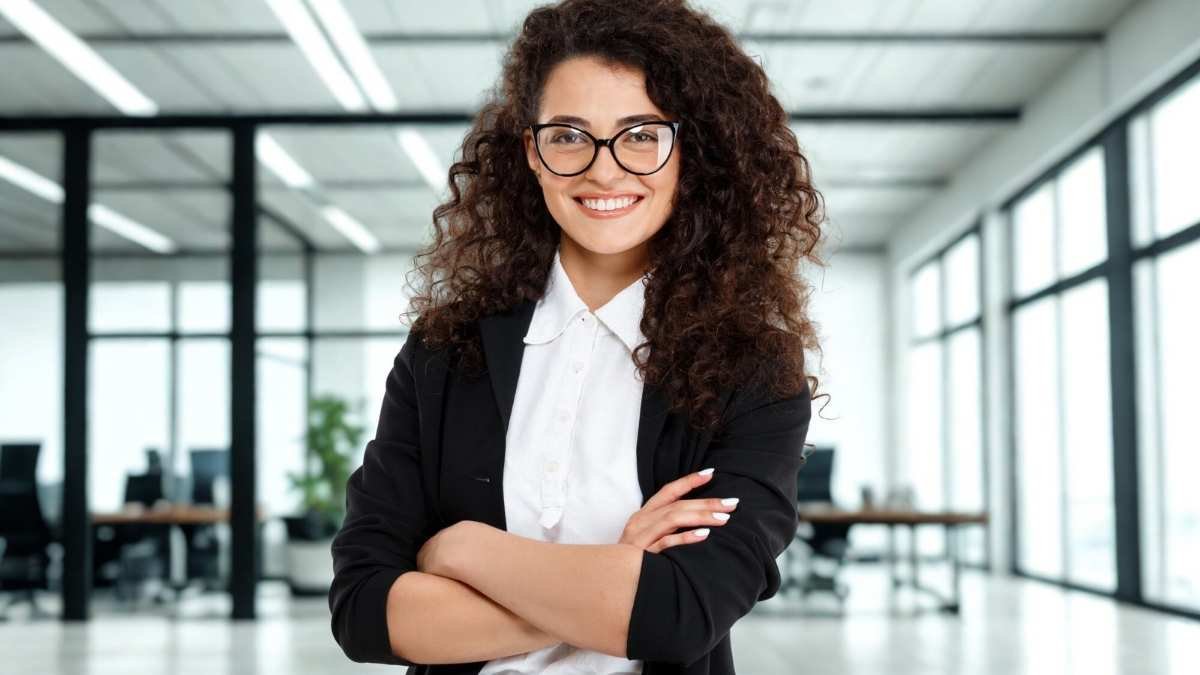 AGEMCAMP abre 2 vagas de estágio em Campinas + CR 1 Mulher jovem com cabelos cacheados e escuros sorri de braços cruzados em um escritório moderno com janelas amplas e iluminação natural ao fundo ela usa óculos de grau com armação escura e está vestida com blazer preto sobre camisa branca o ambiente é corporativo e ao fundo há mesas de trabalho cadeiras e uma planta próxima à parede de vidro - AGEMCAMP abre 2 vagas de estágio em Campinas + CR