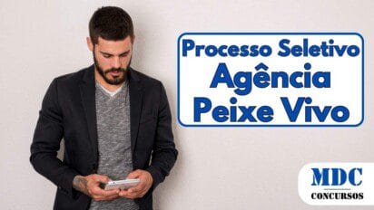 Homem jovem com barba e cabelo curto, vestindo blazer preto sobre camiseta cinza mesclada, segurando um smartphone e olhando para a tela. Ele tem uma tatuagem no braço esquerdo e está encostado em uma parede branca - Agência Peixe Vivo – MG oferece 84 vagas e Salários de até R$ 10,3 mil