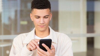Jovem em pé, usando uma jaqueta clara e camiseta branca, olha atentamente para a tela do smartphone que segura com uma mão enquanto digita com a outra, em frente a um prédio com vidros ao fundo. Foto: pch.vector/Freepik