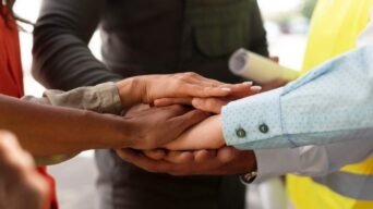 Grupo de pessoas une as mãos em gesto de cooperação, simbolizando diversidade, inclusão e trabalho em equipe entre profissionais de diferentes áreas.