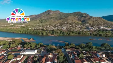 Vista aérea de Almenara com rio, casas, vegetação e montanhas ao fundo, com logomarca da Prefeitura no canto superior esquerdo. Foto: Divulgação/Instituto Nacional do Seguro Social - INSS