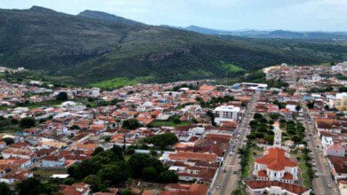 Vista aérea de Alpinópolis-MG com destaque para a igreja matriz, áreas residenciais com telhados vermelhos e montanhas ao fundo.
