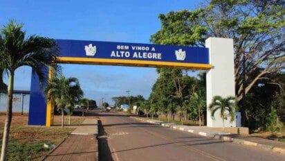 Imagem da entrada da cidade de Alto Alegre, em Roraima, com um grande pórtico azul e branco com a frase "Bem-vindo a Alto Alegre". O pórtico está localizado sobre uma via asfaltada com canteiros laterais, ladeada por palmeiras e vegetação. O céu está limpo e o ambiente transmite uma atmosfera tranquila e organizada, típica de cidades do interior.